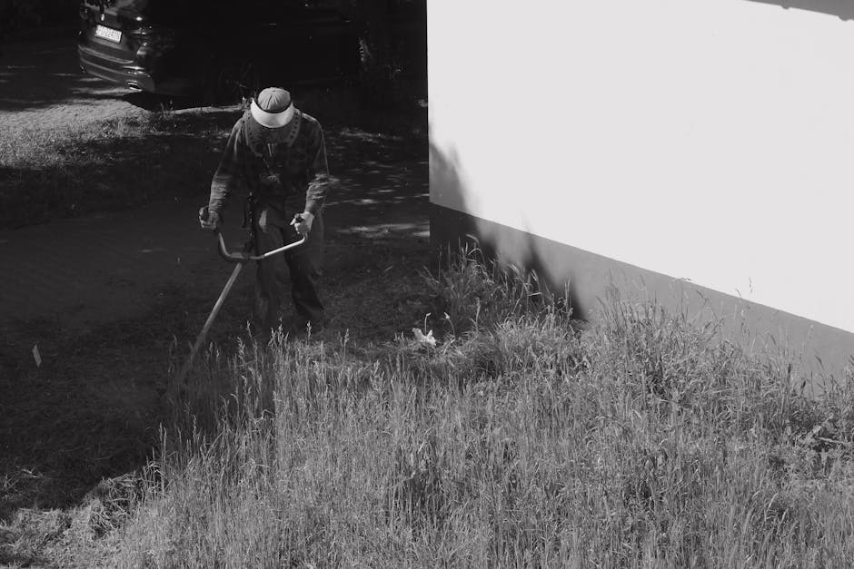 A person engaged in garden maintenance activities, using a long-handled tool to work on a grassy area adjacent to a paved patio. The garden features a mix of tall grass, small plants, and a neatly tri